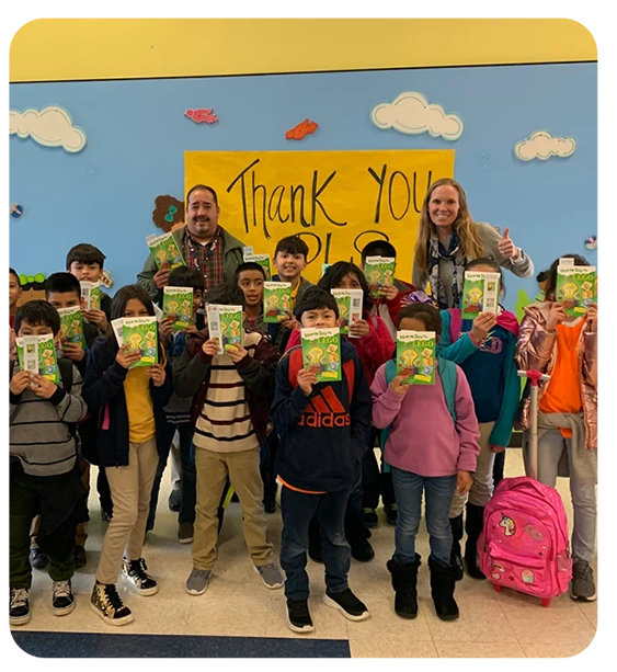 Lowey Bundy Sichol standing with a group of grade school children. Everyone is holding a copy of her book, From an Idea to Lego in front of their faces.