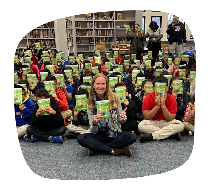 Lowey Bundy Sichol sitting in front of group of grade school children. Everyone is holding a copy of her book, From an Idea to Lego in front of their faces.